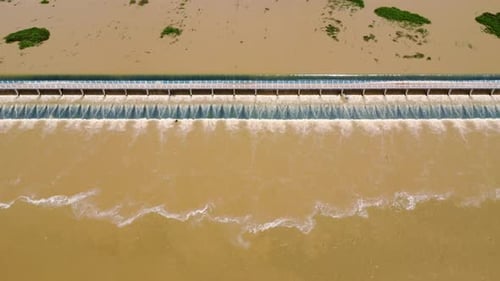 Damm mit schlammigem Wasser, das über seinen Kamm fließt und einen wasserfallähnlichen Effekt erzeugt. - Luftaufnahme