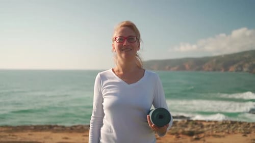 Mature Woman in Glasses Holding Yoga Mat Standing on Beach