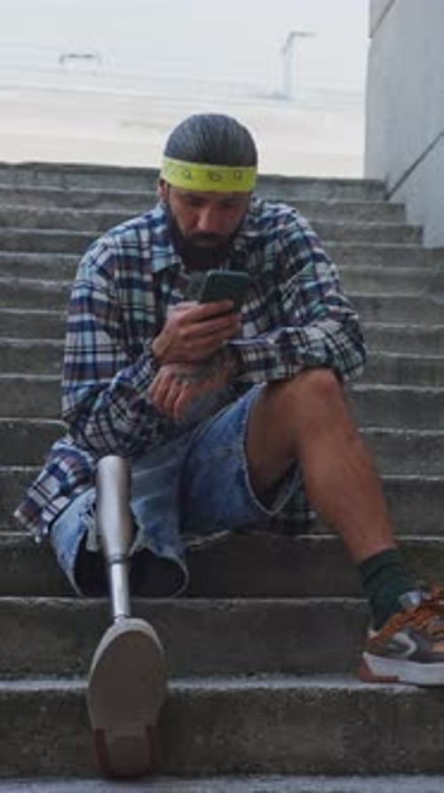 Man Using a Phone While Sitting on Steps with a Prosthetic Leg in an Urban Area