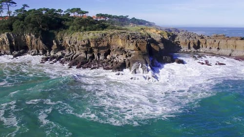 Aerial view of the waves crashing on the rocky cliff in Cantabria, Spain during a sunny day.