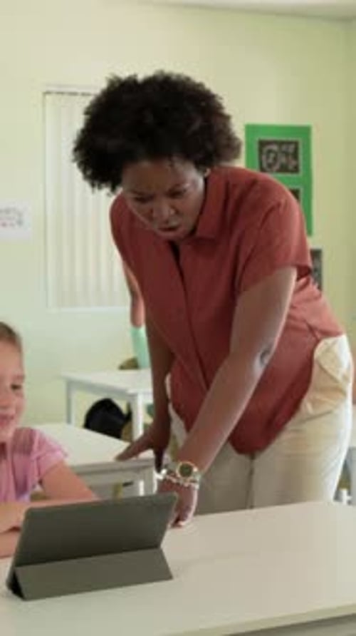 Vertical video: Entering aisle teacher in rust shirt leaning over tablet and laptop, helping pupils