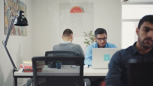 A Man in a Blue Shirt in His Glasses Works in a Computer at the Office Around Colleague Business