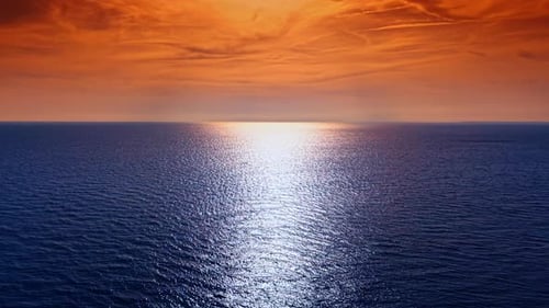 Endless blue waterscape with sparkling light path. Cloudy orange sky is above the horizon.
