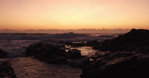 Slow motion shot of ocean waves breaking on rocks, golden orange sunrise