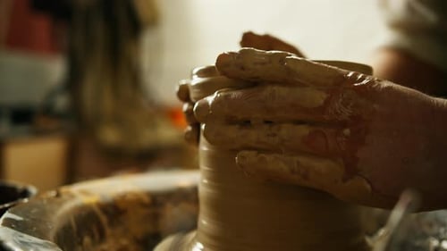 Close-up of potter's hands covered with clay making beautiful vase on throwing wheel in pottery