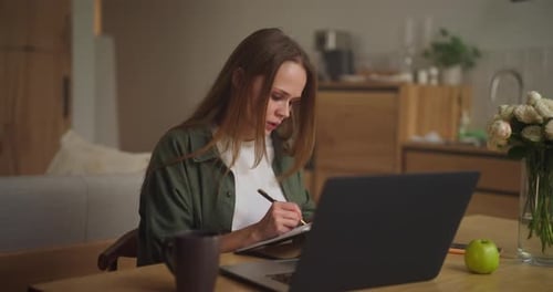 Une fille blonde travaille et étudie à distance sur un ordinateur portable, écrit des plans importants sur un bloc-notes et