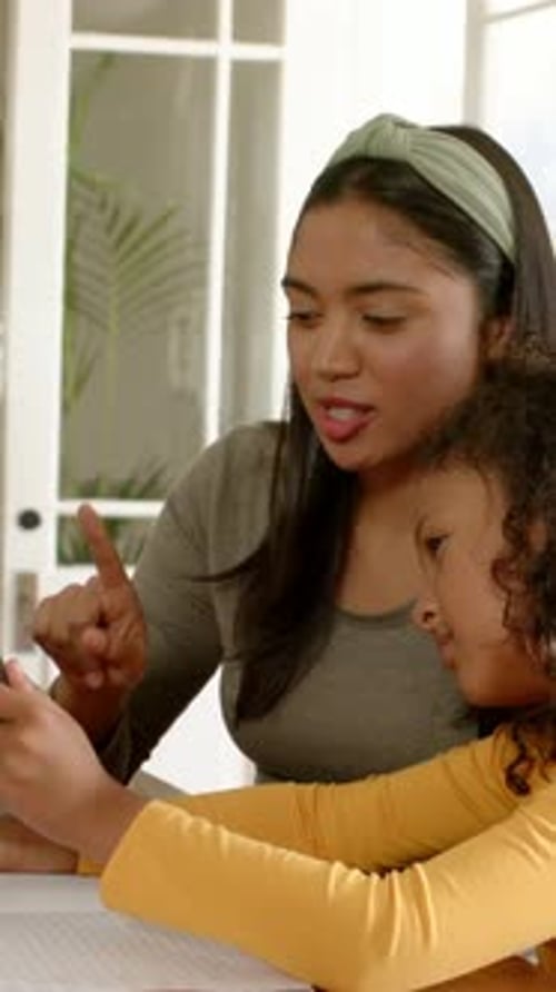 Vertical video: Leaning woman pointing, teaching girl using tablet at home with notebook pencilcase