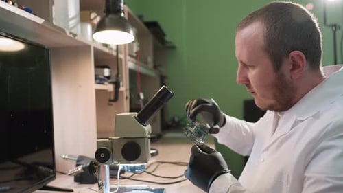 Technician Examining Circuit Board Under Microscope in WellLit Electronics Lab