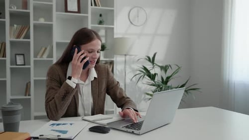 Focused Businesswoman Speaking on the Smartphone While Sitting at Home Office and Using the Laptop
