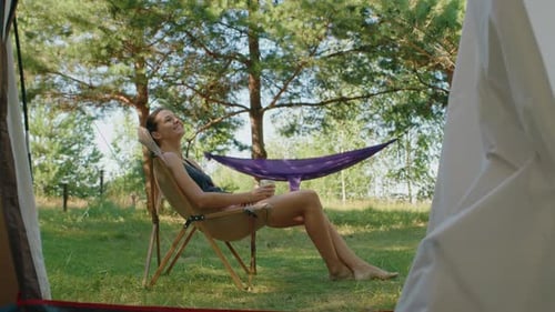 Young Woman Enjoying a Cup of Coffee While Sitting on a Camping Chair Near a Hammock in a Forest