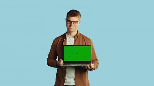 Young Smiling Caucasian Man in Glasses and Casual Clothes Showing a Laptop with a Mockup Green