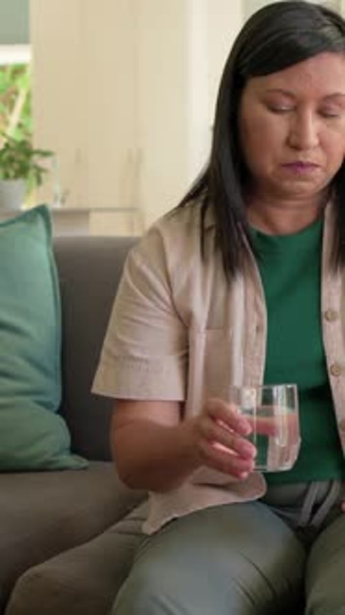 Vertical video: Leaning mature woman green top grasping glass on sofa placing pill to swallow