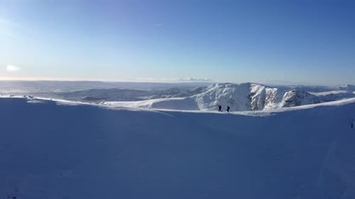 Couple walking a snowy peak of a great valley in the sunshine. Aerial dolly out