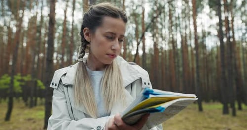 Blonde Beautiful Woman with Braids Wearing Sportswear Standing in the Middle of Forest Holding Map