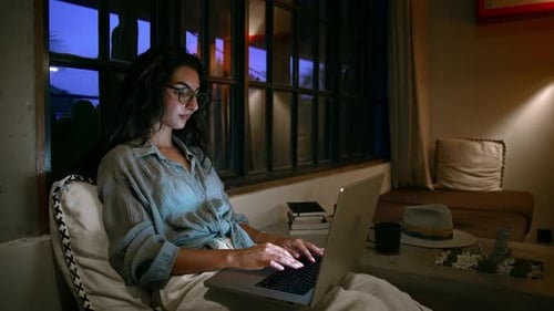 Young Woman in Home Office Looks at Laptop Screen and Works Typing on Keyboard