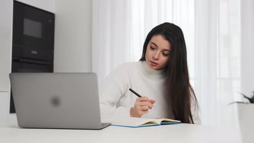 Woman Writing in Notebook With Laptop at Home