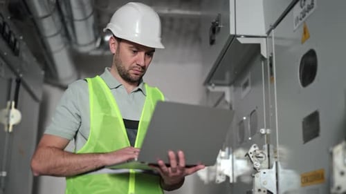 Electrical Engineer Using Laptop Next to Control Panel in Factory
