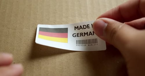 Hands applying MADE IN GERMANY flag label on a shipping box with product premium quality barcode. Ma