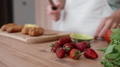 A woman is cooking behind the wooden table. She takes strawberries and starts cutting them