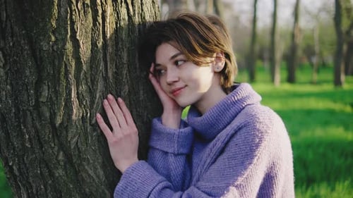 Young Woman Enjoys Nature While Resting Against a Tree in a Green Park During a Sunny Day in