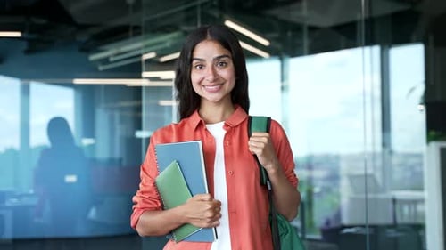 Portrait of smiling young female student standing in modern university hallway holding books