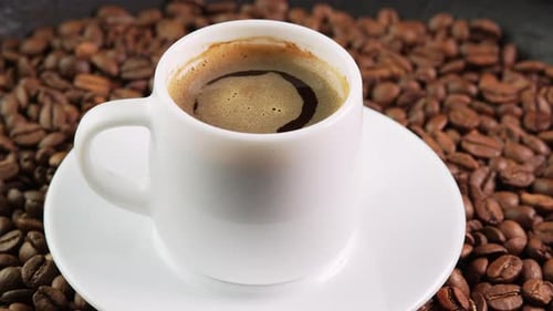 White coffee cup with foam black coffee rotates on a saucer among roasted beans background close
