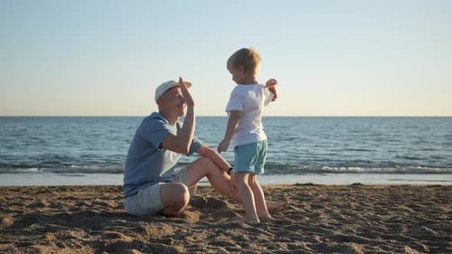 A Little Boy Gives His Dad a High Five Sitting on the Beach During Sunset