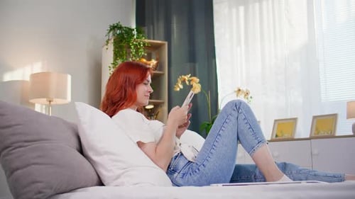 Freelancer Young Woman Works Remotely Using a Mobile Phone on Internet While Lying on Bed at Home