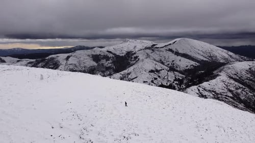 Solitary hiker climbing white snowed capped mountain, stops for a break, aerial
