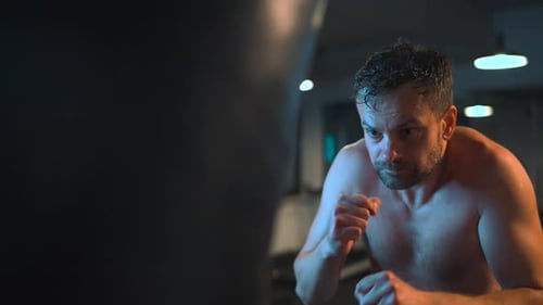 Alone Boxer is Working Out Blows on a Punching Bag in a Gym in Evening
