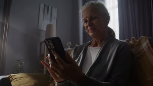 Happy Retired Woman Uses Mobile Phone Resting on Cozy Sofa
