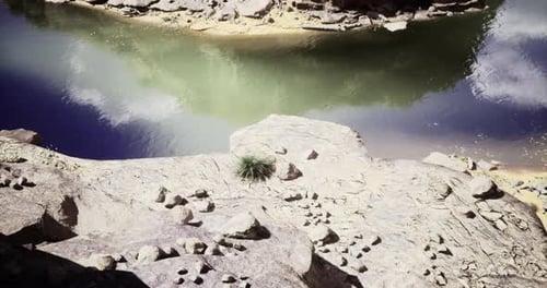 Rocky Shoreline Beside Tranquil River Reflecting Clouds in Clear Sky