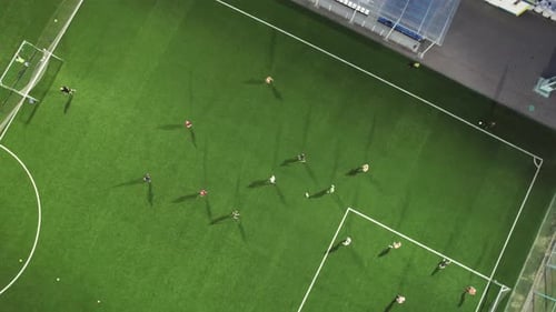 Aerial view: Football players play soccer at night on a green field lit by artificial lights.