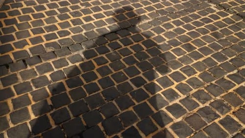 Ein Mann wirft seinen eigenen Schatten, während er auf einem gemauerten Straßenpflaster steht, während andere Leute an Casti vorbeikommen