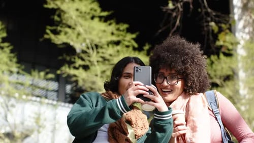 Amigas diversas estendendo o smartphone para selfie, segurando café e andando para fora do prédio