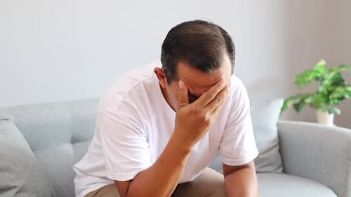 Stressed Asian man sitting on sofa with hands covering face showing emotional struggle anxiety and