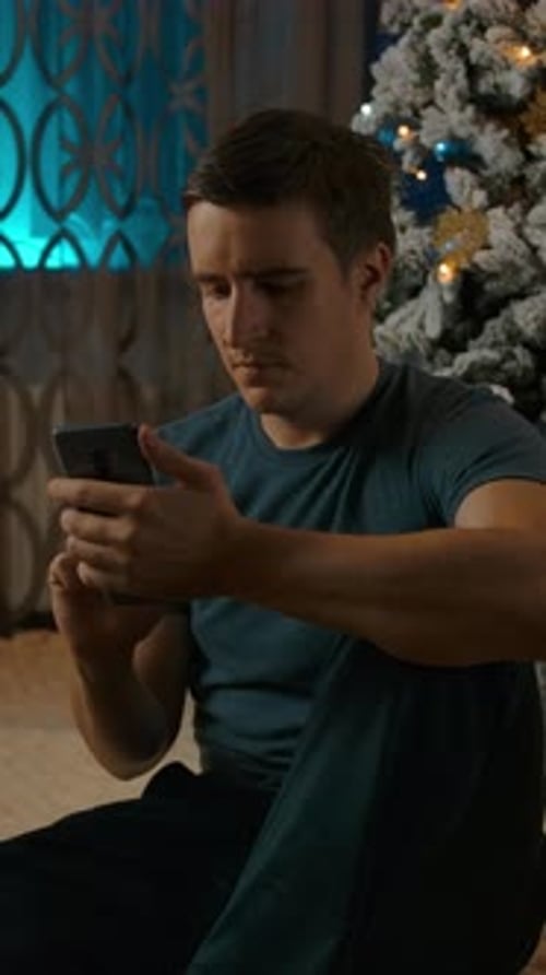 Vertical of Young Caucasian Man Using Smartphone Sitting Near Cozy Christmas Tree Space