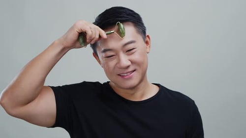 Studio Portrait of Young Chinese Handsome Man Using Jade Roller Massager for Antiwrinkle Beauty