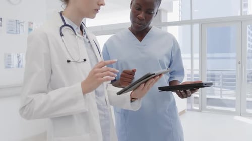 Diverse female doctor and nurse using tablets and discussing in hospital corridor, in slow motion