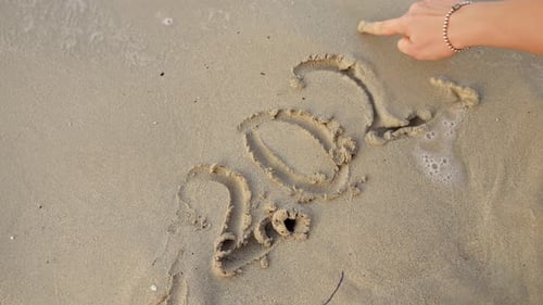 Closeup of female hand using finger to write 2023 year on wet sand at ocean beach.