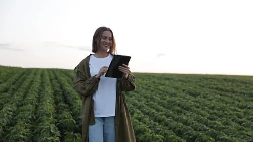 Smiling female agronomist walking at soybean plantation with digital tablet, examines plants and doe