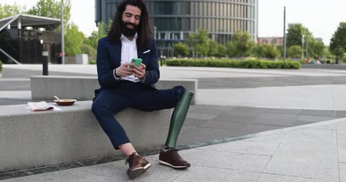 Business Man With Prosthetic Leg Using Mobile Phone Outdoor From Office Building - Job And Insurance
