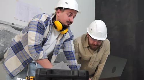 Two Engineers in Helmets are Discussing Documentation on a Laptop