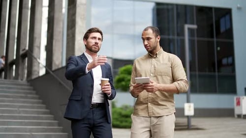 Two professional businessmen communicate while walking outside near office building