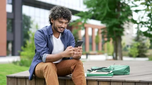 Male student uses a mobile phone while sitting on bench in campus space near university building.