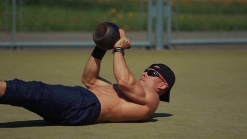Young Man Training on Outdoor Sports Field and Performs Twohanded Kettlebell Press While Lying on