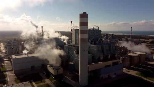 Factory complex with tall chimney and steam rising into atmosphere, aerial orbit view