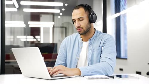 Focused African American IT specialist in wireless headphones works on a laptop sitting