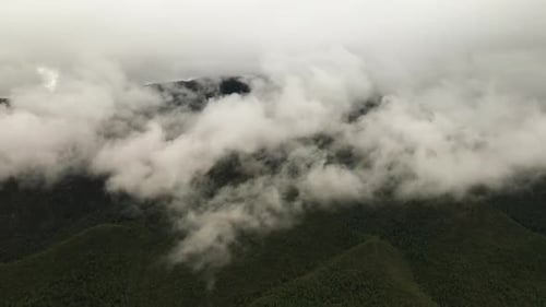 Drone shot of green mountains surrounded by dramatic grey clouds. The camera glides beside the peaks