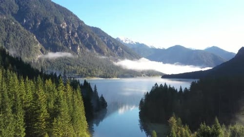 Aerial shot of a big mountain lake surrounded by forests and covered in clouds during sunrise